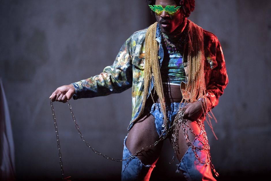 emeka ene | in Doom emeka ene steht vor dunkelgrauem Hintergrund. Er trägt an der Leiste ausgeschnittene Jeanshosen, eine bunt gefärbte Jeansjacke und viele Perlenschnüre um den Hals. In den Händen hält er eine lange silberne Kette.