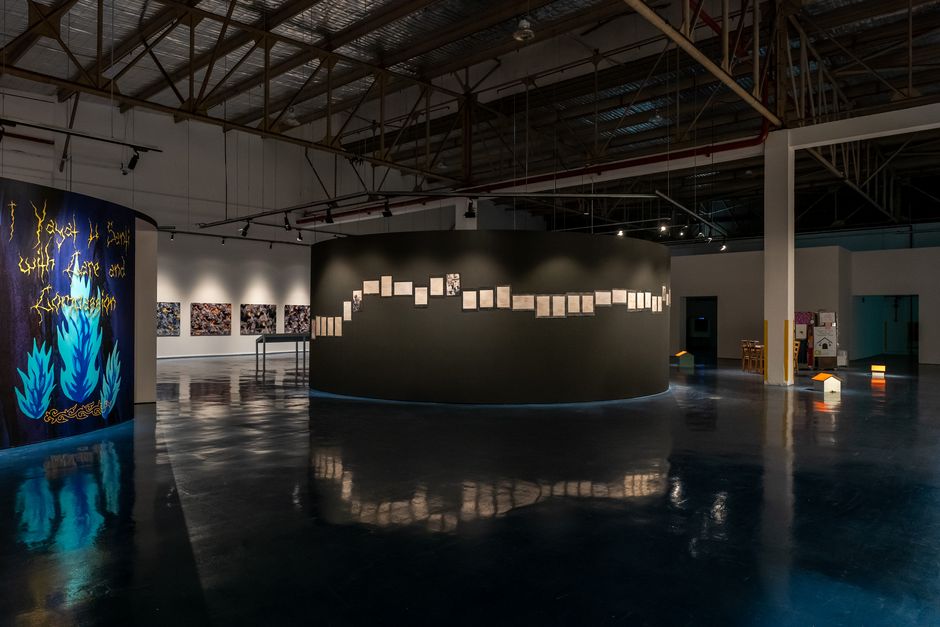 Kiraṇ Kumār, SIX UNEASY FRAGMENTS (EXACTLY) ABOUT THE NATURAL AND SPIRITUAL Ein Ausstellungsraum mit einer gerundeten dunkelgrauen Wand rechts, an der Bilderrahmen hängen, und einer Wand mit blauen Flammen links. Im Hintergrund hängen Bilder an einer weißen Wand.