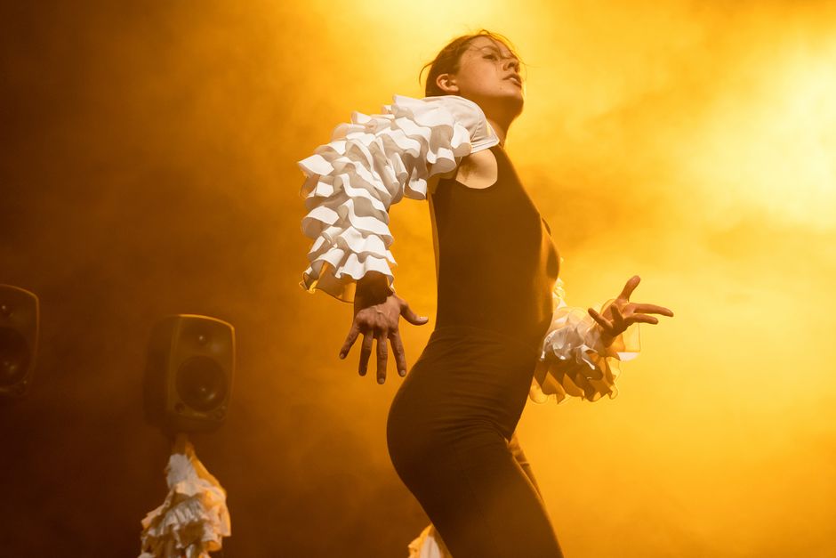 Luisa Fernanda Alfonso, Masterpiece Eine Person mit schwarzem Bodysuite und weißen Ärmeln in einer Bewegung. Der Hintergrund ist gelb erleuchtet.