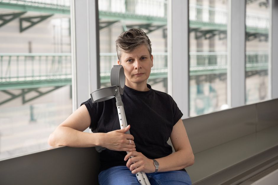 Ein Porträt von Claire. Claire hat kurzes dunkelbraunes Haar mit grauen Flecken an den Schläfen und blaue Augen. Sie trägt ein schwarzes T-Shirt. Mit ihr sind zwei graue, altmodische britische Ellbogenkrücken. // Englisch: A portrait of Claire. Claire has short dark brown hair with grey patches at her temples, and blue eyes. She is wearing a black sthirt. With her, are 2 elbow crutches - they are grey and of a quite old fashioned UK design.