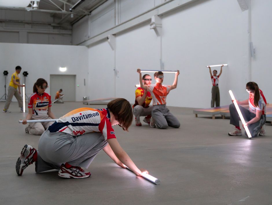 Hannah Schillinger | runners Acht Personen in Sportklamotten befinden sich sitzend oder stehend in einer grauen Halle und halten Leuchtende Röhren in den Händen.